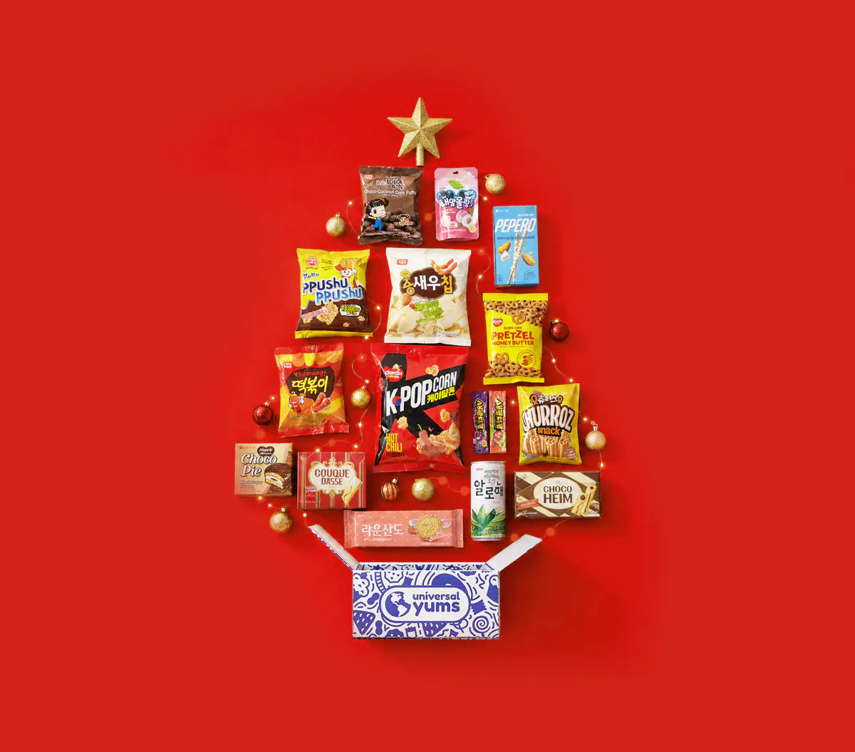 Korean snacks and gold ornaments arranged in the shape of a holiday tree, topped with a gold star, above an open Universal Yums box.
