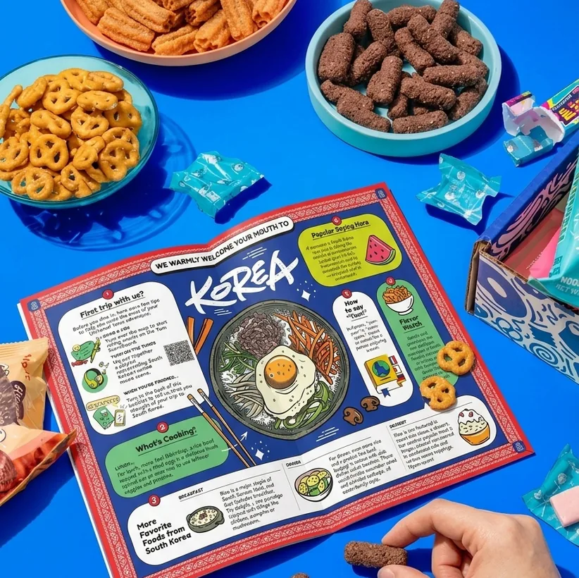 The Korea tasting mat on a blue table with bowls of snacks, and a hand picking up a chocolate stick.