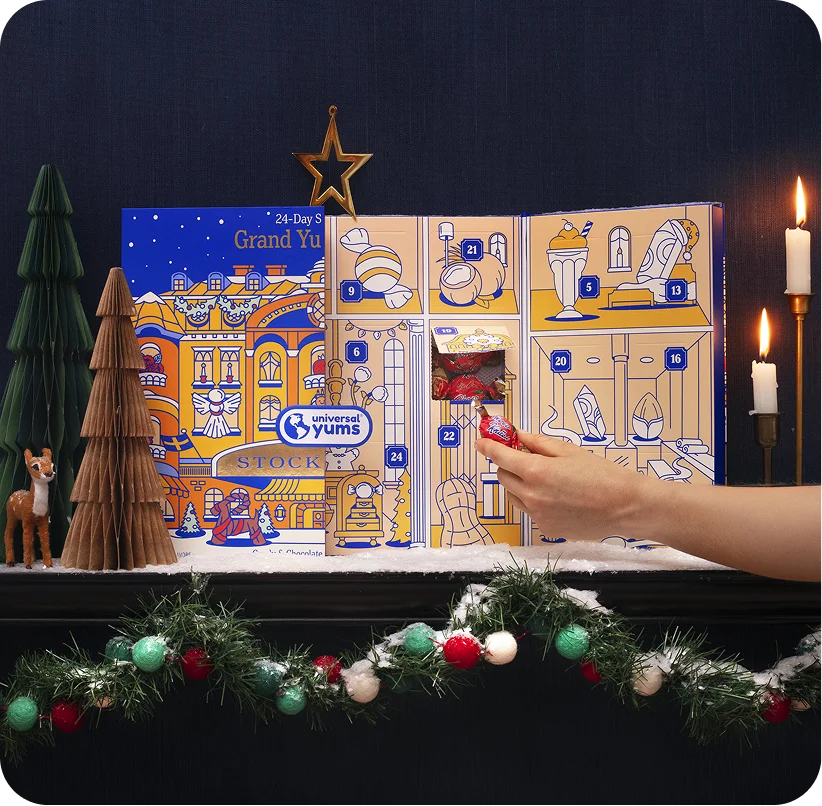 A hand opening a door of the Grand Yums Hotel advent calendar standing between paper trees and a garland.